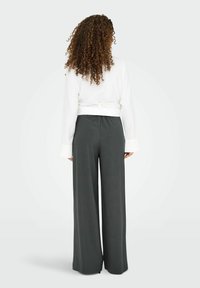 Femme aux cheveux bouclés portant un haut blanc à manches longues et un pantalon large taille haute gris foncé, debout de dos sur un fond uni.