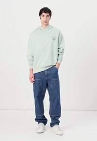 Hellgrünes Hoodie mit einem kleinen Grafik, kombiniert mit lockeren blauen Denim-Jeans. Die Textur ist weich und der Schnitt ist lässig. Weiße Sneakers runden den Look ab.