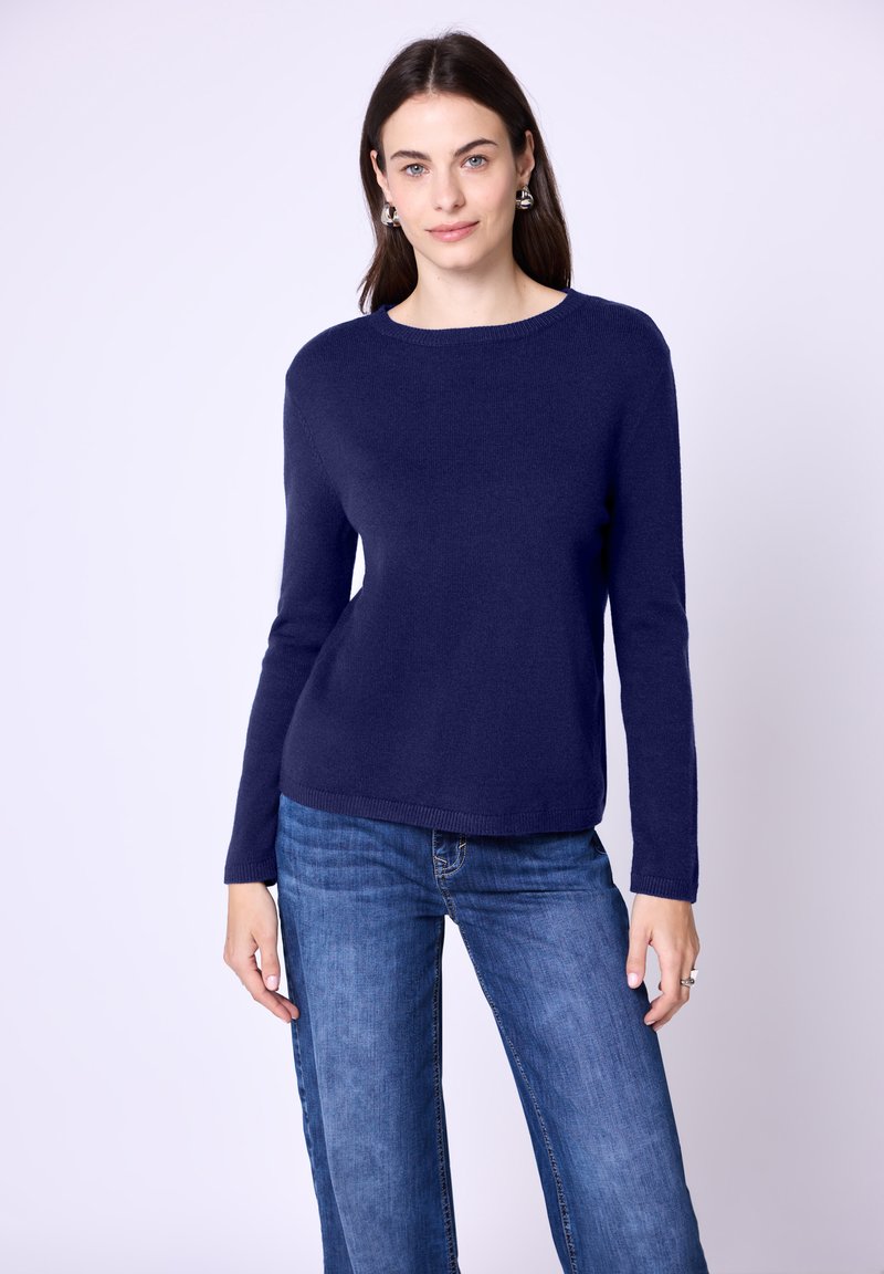 Frau mit langen dunklen Haaren, die silberne Ohrringe, einen dunkelblauen langärmeligen Pullover und blaue Jeans trägt, vor einem hellen Hintergrund steht.
