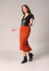 T-shirt noir à col en V avec le texte en paillettes "LOVE", accompagné d'une jupe midi en velours côtelé orange et de bottines noires, se tenant devant un fond neutre.