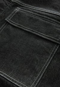 Tessuto in denim nero con una tasca applicata e cuciture bianche a contrasto. La texture del materiale appare leggermente sbiadita con una finitura liscia.