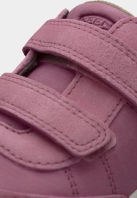 Chaussure avec bride en velcro rose et texture douce ressemblant à du suède, présentant des coutures contrastées et un bout arrondi, adaptée pour un usage décontracté.