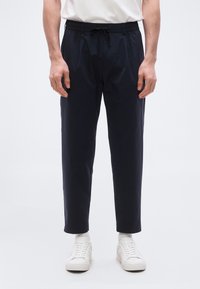 Άνδρας που φοράει μαύρο παντελόνι με κορδόνι, άσπρες κάλτσες και λευκά αθλητικά παπούτσια, στέκεται μπροστά σε απλό φωτεινό φόντο.