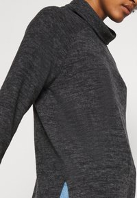Personne portant un pull à col roulé à manches longues gris foncé, associé à un jean bleu clair, vue partielle de profil.