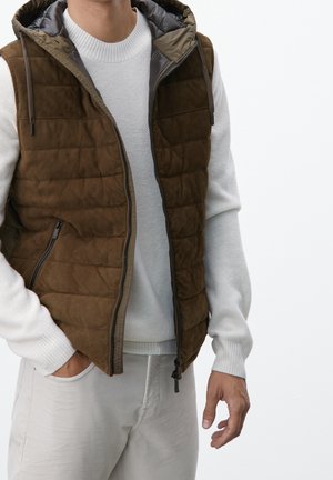 Uomo con gilet trapuntato marrone sopra maglione bianco di maglia, con una mano in tasca e pantaloni bianchi su sfondo bianco.