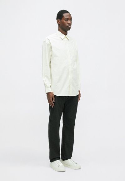 Jasno beżowa koszula zapinana na guziki z długimi rękawami, w zestawie z czarnymi spodniami i białymi trampkami. Prosta, gładka tekstura, minimalistyczny design.