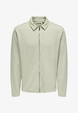 Hele roheline tõmblukuga jope, millel on terav kaelus ja mis on valmistatud tekstuursetest kangast. Omab pikki varrukaid ja sujuvat tõmbluku avamist ees.