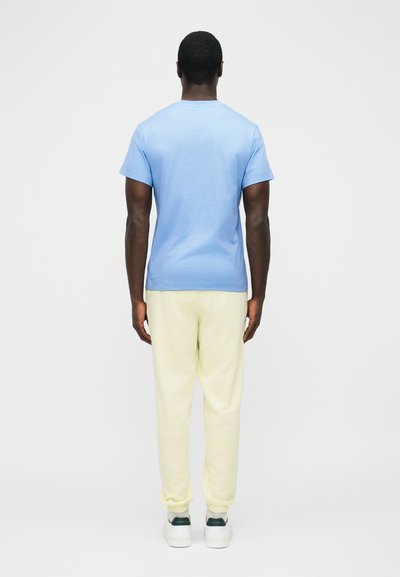 Άνδρας που στέκεται πλάτη φορώντας μπλε γαλάζιο μπλουζάκι, κρεμ φόρμα και λευκά αθλητικά παπούτσια σε απλό λευκό φόντο.