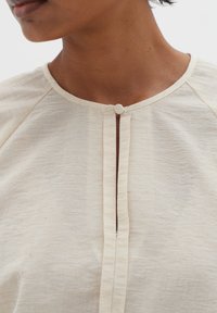Primo piano di una persona che indossa una blusa beige con texture, dotata di scollatura rotonda e di un solo bottone rivestito in tessuto al centro in alto.