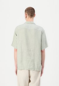 Šviesiai žalia trumpomis rankovėmis mar shirt iš tekstūrinio lininio audinio, su sagų apykakle ir tiesiu apdailu, rodoma iš nugaros.