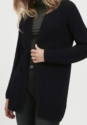 Femme portant un cardigan noir texturé à ouverture frontale avec des poches, par-dessus un col roulé sombre et un pantalon noir.