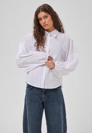 Vrouw met lang golvend haar draagt een los wit overhemd met brede mouwen en een blauwe jeans, staand tegen een effen lichte achtergrond.
