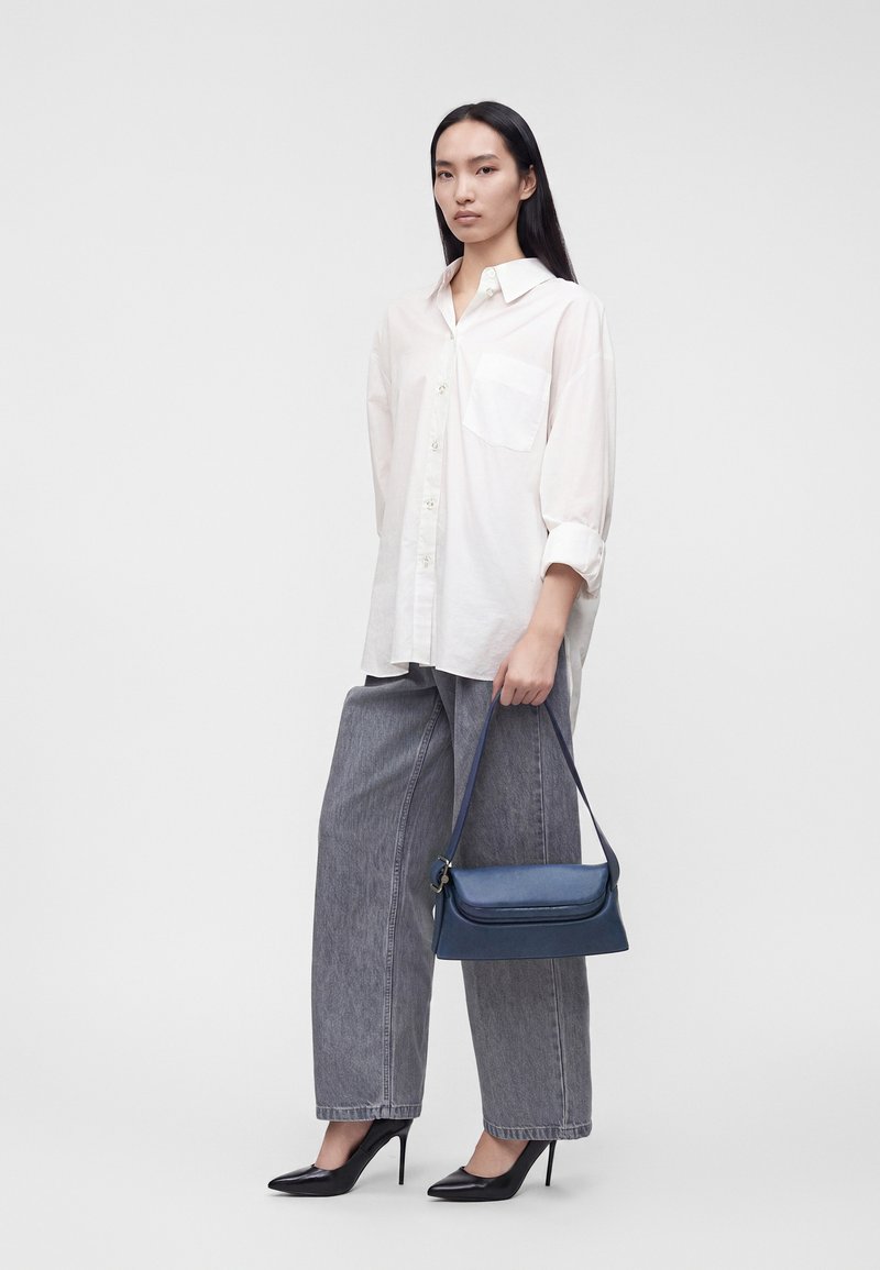 Femme aux longs cheveux foncés portant une chemise blanche boutonnée, un jean gris à jambes larges, des talons hauts noirs, tenant un sac à main bleu marine.