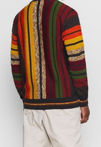 Homme portant un pull en tricot rayé multicolore avec des motifs verticaux rouges, jaunes, verts, oranges et bruns, assorti de pantalons beige clair.