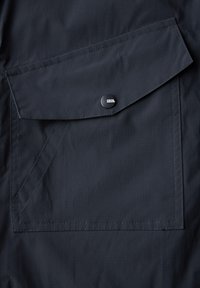 Tejido ligero de color azul marino con un bolsillo con solapa que presenta un botón negro y el logotipo "CECIL". Textura suave y detalles de costura reforzada.