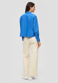 Kurze blaue Button-up-Bluse mit Kragen, kombiniert mit hellbeigen weiter geschnittenen Hosen und weißen Sneakern. Glattes Material und lockere Passform.
