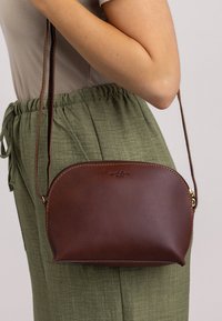 Ruskea nahkainen olkalaukku pyöreällä muodolla ja kullatuilla yksityiskohdilla. Hihna on säädettävä. Esitelty vaaleaa taustaa vasten.