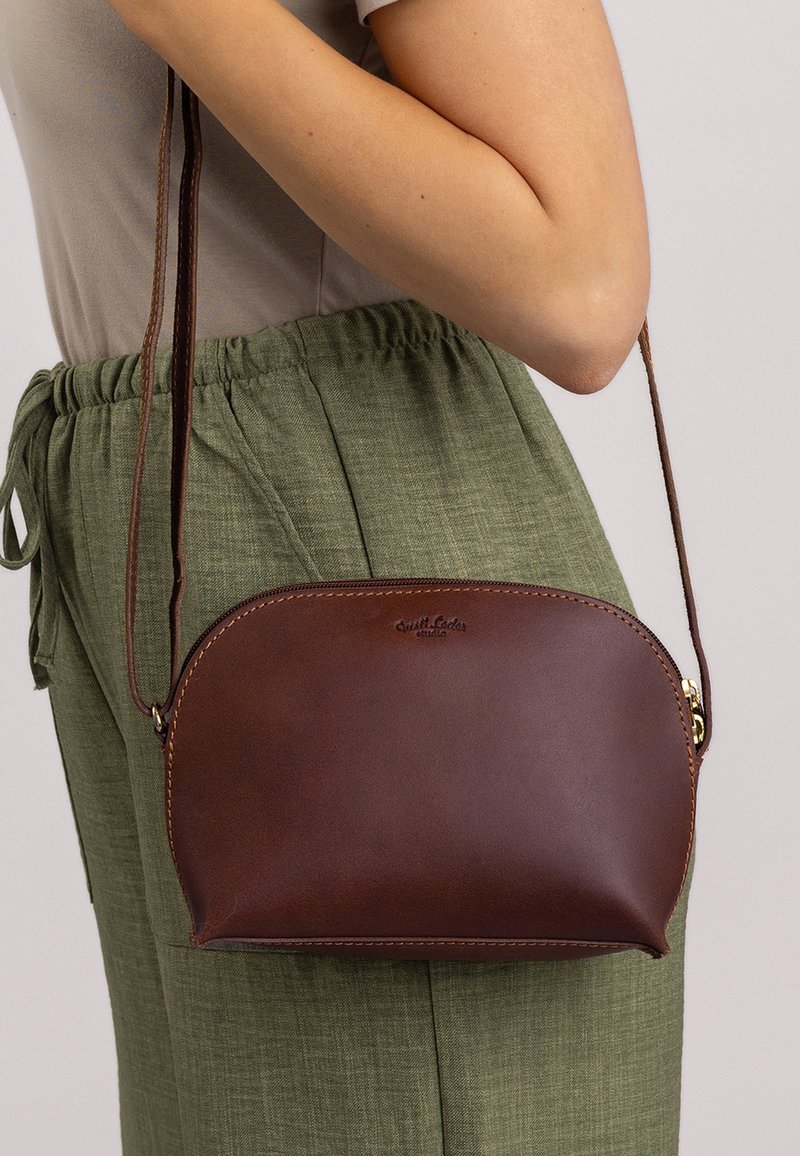 Ruskea nahkainen olkalaukku pyöreällä muodolla ja kullatuilla yksityiskohdilla. Hihna on säädettävä. Esitelty vaaleaa taustaa vasten.