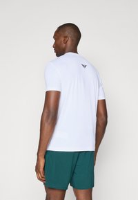 Valge sportlik T-särk lühikeste varrukatega, millel on tekstuuriga kangas ja must logo selja ülaosas. Seljas on rohelised lühikesed püksid.