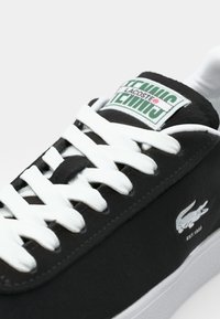 Sneaker noire Lacoste avec lacets blancs, semelle blanche, logo vert et blanc sur la languette et emblème crocodile sur le côté.