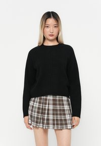 Maglione nero a maglia con scollo rotondo, abbinato a una gonna corta a quadri nei toni del marrone e del bianco, su uno sfondo neutro.