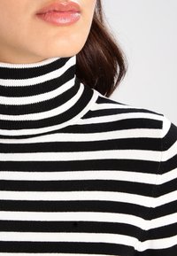 Musta-valkoraidallinen pooloneule, jossa on resorikuvio ja istuva malli, korostaen vuorottelevia paksuja ja ohuita raitoja.