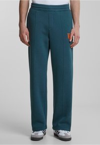 Pantalones de chándal en color verde azulado con un ajuste relajado, cintura elástica, bolsillos laterales y un logo naranja en el muslo izquierdo. Fabricados con un tejido suave.