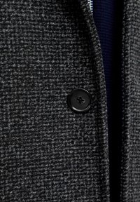 Grå struktureret stoffrakke med en sort knap. Materialet har et vævet mønster, med en glat overflade synlig ved kraven.