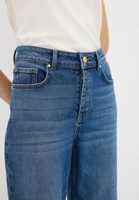 Jeans a vita alta di un blu medio, con elementi in metallo dorato, design classico a cinque tasche e leggere sfumature per aggiungere texture.