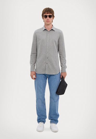 Gray long-sleeve shirt, blue jeans, white sneakers. The person holds a small black bag. Sunglasses complete the outfit.