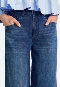 Sötétkék, széles szárú farmernadrág sima textúrával. Két első zsebbel és klasszikus gombos derékzárral rendelkezik.