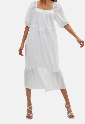 Kobieta ubrana w białą sukienkę midi z bufkami na rękawach długości do łokcia, kwadratowym dekoltem, wielowarstwową spódnicą i białymi sandałami na wysokim obcasie z paskami.