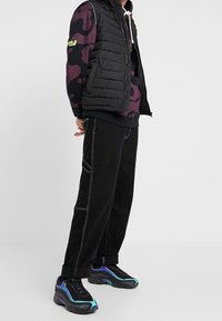 Černá péřová vesta vrstvená přes černou mikinu s vzorovými akcenty, spárována s volnými černými džíny a černými botami s tyrkysovými a fialovými detaily.