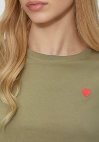 Zielono-oliwkowy bawełniany t-shirt z okrągłym dekoltem i krótkimi rękawami, z małym haftem czerwonego serca po lewej stronie klatki piersiowej.