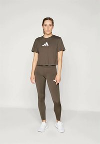 Oliivroheline lühike t-särk valge logoga, millele on sobitatud elegantsed retuusid. Mudel kannab valget sportjalatsit ja seisab neutraalse tausta ees.