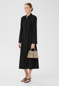 Abito lungo a maniche lunghe nero con bottoni sul davanti e pence; borsa a tracolla beige con stampa serpente testurizzata; infradito nere; sfondo bianco puro.