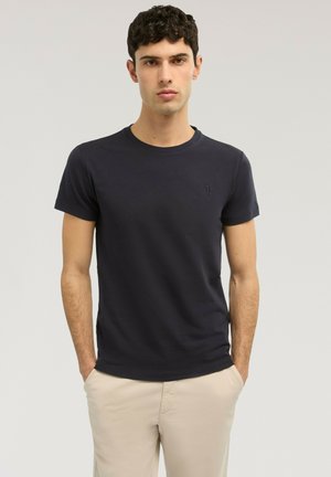 Junger Mann trägt ein schlichtes schwarzes T-Shirt und beige Hosen, steht mit den Händen in den Taschen vor einem neutralen Hintergrund.