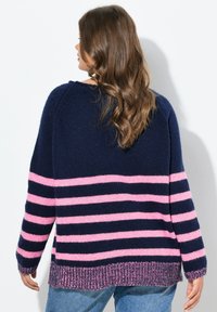 Vrouw met lang golvend haar, gekleed in een marineblauwe trui met roze horizontale strepen, in combinatie met blauwe jeans, met de rug naar de camera gekeerd.