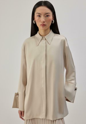 Beżowa jedwabna bluzka z kołnierzykiem, długimi rękawami i oversize'owymi mankietami ozdobionymi guzikami. Gładka faktura, luźny krój.