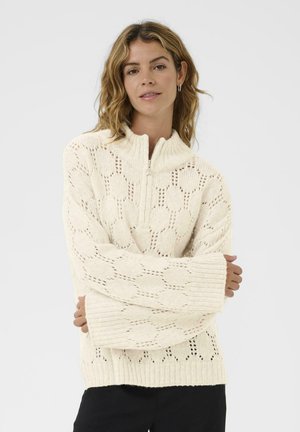 Frau mit welligem Haar trägt einen cremefarbenen Strickpullover mit einem Sechseckmuster und einem Viertelreißverschluss-Kragen. Sie steht mit verschränkten Armen vor einem weißen Hintergrund.
