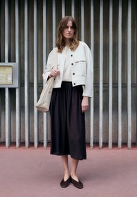 Woman standing in front of vertical concrete bars, wearing a white jacket, beige top, black midi skirt, brown shoes, and carrying a beige tote bag.