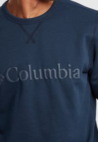Felpa blu navy con colletto e polsini a coste, con un logo "Columbia" in rilievo sulla parte anteriore. Tessuto morbido.