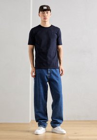 Tricou scurt cu mâneci, pantaloni din denim albastru lejeri, adidași albi, pălărie maro cu patch logo. Design simplu, detalii de cusătură vizibile.