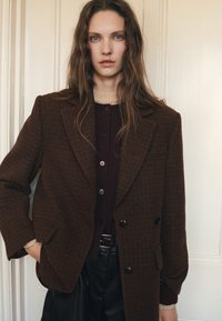 Ruutupainotteinen ruskea kaksirivinen bleiseri, jossa on jämäkät olkapäät, päällä tumman värinen neuletakki, yhdistettynä mustiin korkeavyötäröisiin housuihin.