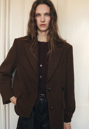 Blazer doppiopetto a quadri marroni con spalle strutturate, indossato sopra un cardigan scuro, abbinato a pantaloni neri a vita alta.