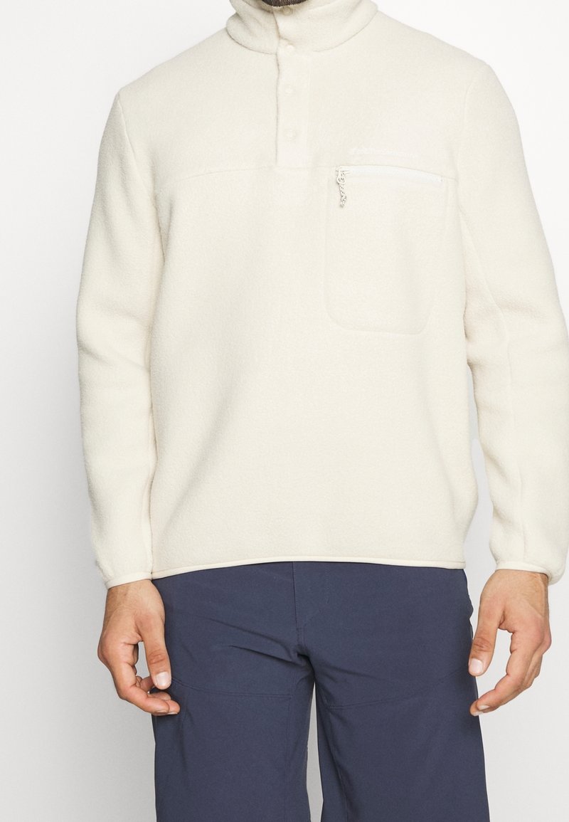 Homme portant un pull polaire blanc avec une poche poitrine zippée et un pantalon bleu marine, visible du cou jusqu'au milieu de la cuisse.