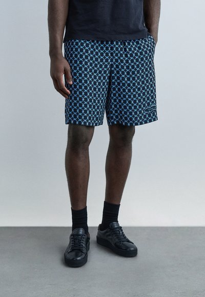 Homme portant un short bleu à motifs, des chaussettes noires et des baskets noires, debout avec une main dans la poche sur un fond uni.