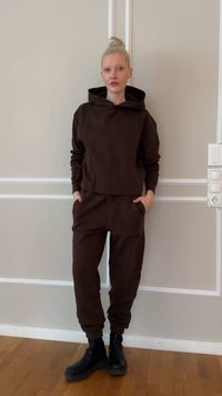 Νέος με ξανθά μαλλιά σε κότσο που φοράει σκούφαν καφέ φούτερ με κουκούλα και αντίστοιχο παντελόνι φόρμας με μαύρες μπότες, στέκεται μέσα σε εσωτερικό χώρο.