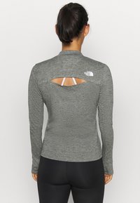 Šedý dlouhý rukáv s vysokým límcem, s výřezem na zádech a bílým logem North Face na horní části zad. Hladká textura.