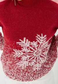 Punainen neulepaita, jossa on korkea kaulus. Paidan alareunassa on valkoinen lumihiutalemalli, ja se on pehmeä ja pörröinen.